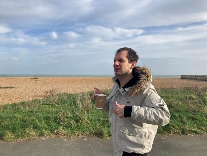 A person near a beach wearing a jacket and holding a reusable cup of tea