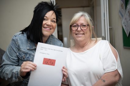Two women smiling for a photo, with one holding a piece of paper