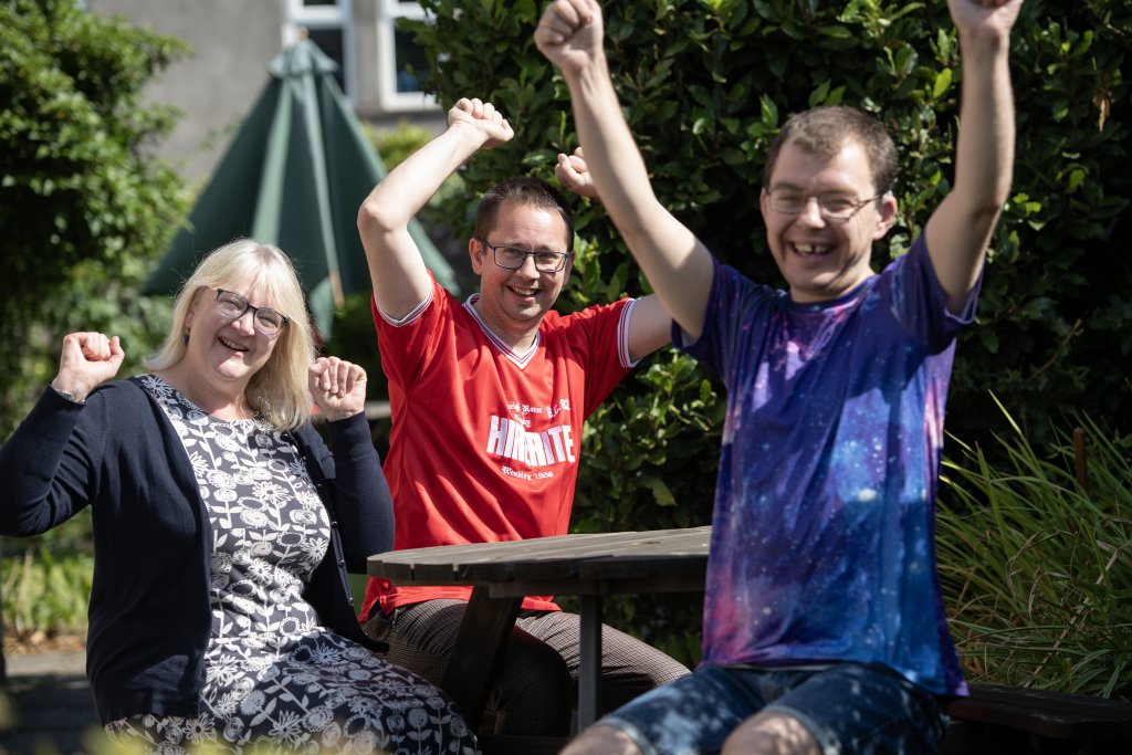 Three people with their arms in the air sat on a bench posing for a photo