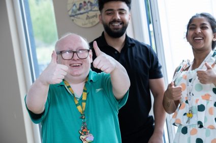 Three people, two men and a woman, smiling at the camera in a living room environment with their thumbs up