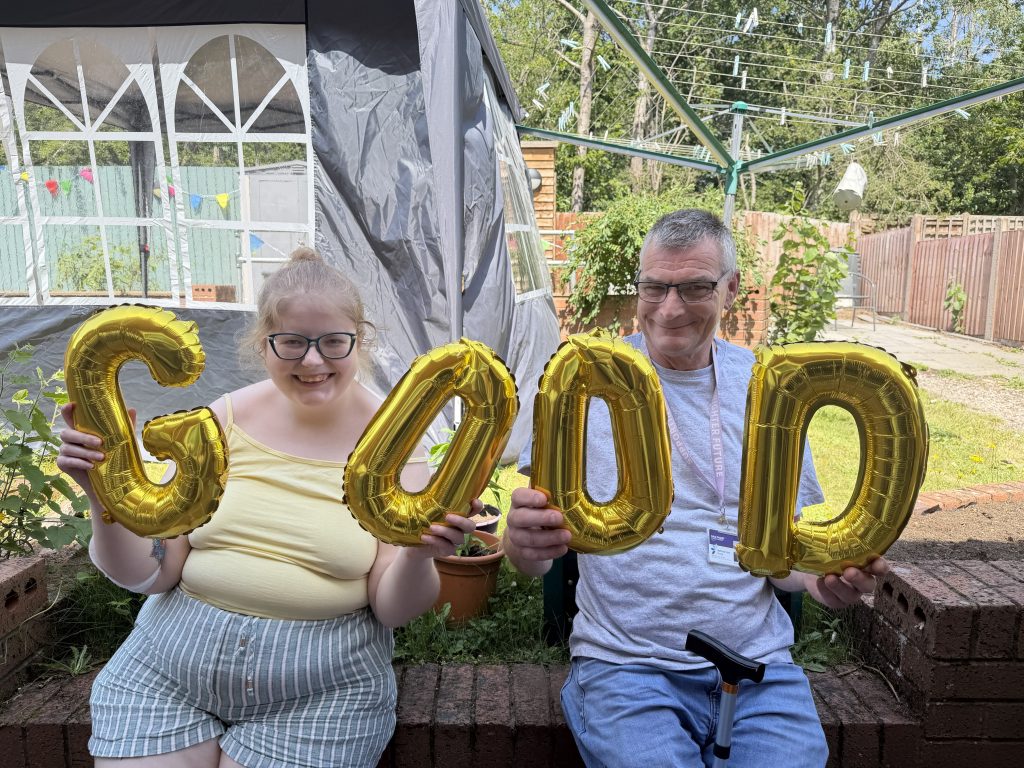 Two people sat on a wall holding golden balloons that spell out GOOD
