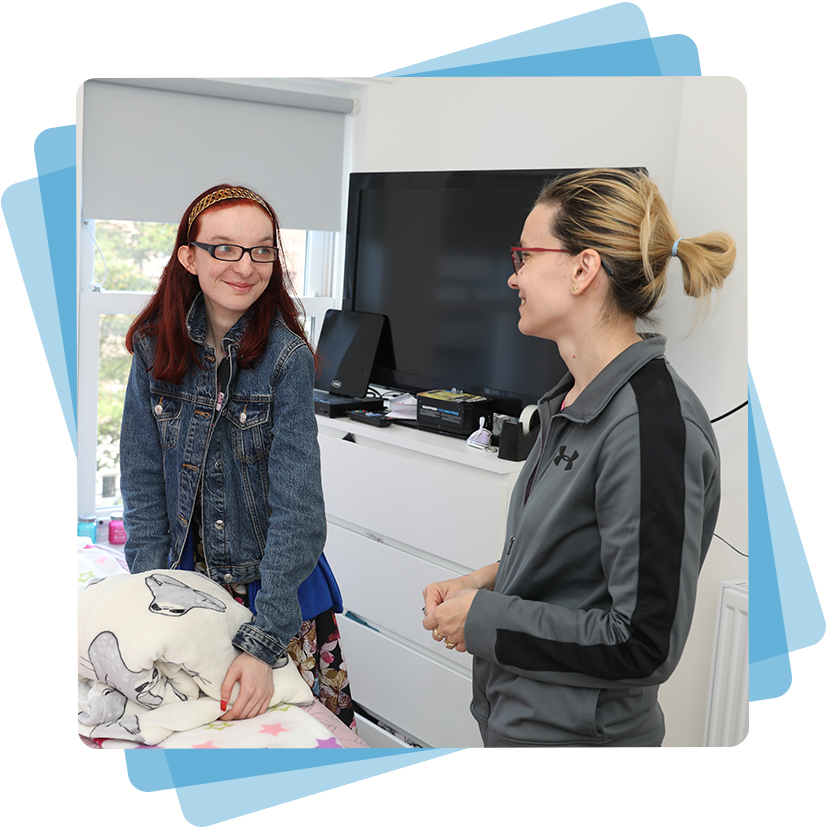 Two women stood in a bedroom talking to each other. The image has a blue box around it.