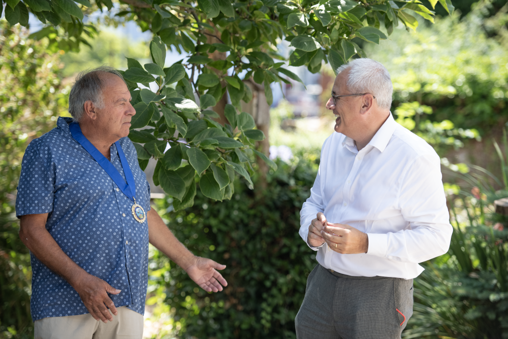 Two men engaged in conversation outdoors surrounded by greenery and sunlight. One man wears a blue patterned shirt with a medal around his neck, while the other wears a white shirt and grey trousers