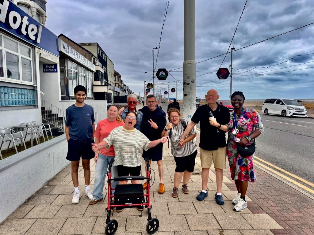 A group of eight adults posing on a seaside sidewalk outside a hotel. Cloudy sky, tram wires, oceanfront road, colorful hanging street decorations, and a red rollator walker highlight an accessible community outing.