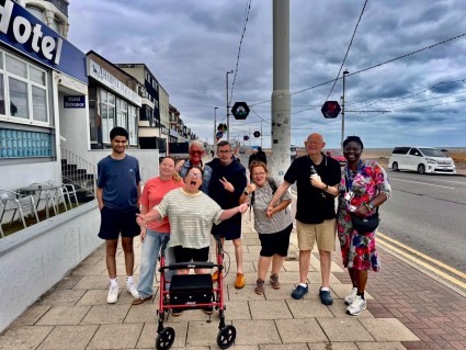 A group of eight adults posing on a seaside sidewalk outside a hotel. Cloudy sky, tram wires, oceanfront road, colorful hanging street decorations, and a red rollator walker highlight an accessible community outing.