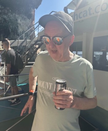 An older man wearing a cap, sunglasses, and “Mount Pleasant” T-shirt while holding a canned drink on vacation