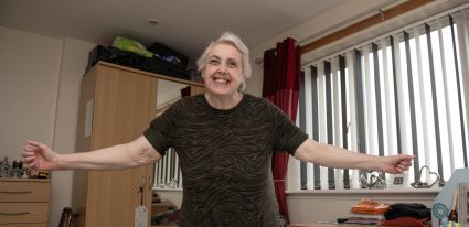 A smiling older woman standing with arms outstretched in bedroom, conveying warmth and joy. Room includes wardrobe, desk, vertical blinds, red curtains, folded pink towels, and floral bedspread.