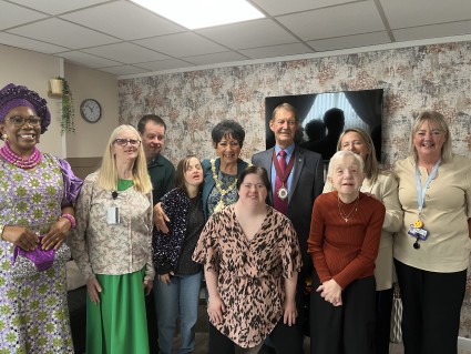 Councillor Joan Bell and her husband, Consort Dennis Bell, posing for a photo with a group of diverse people. They appear to be in a living room environment.