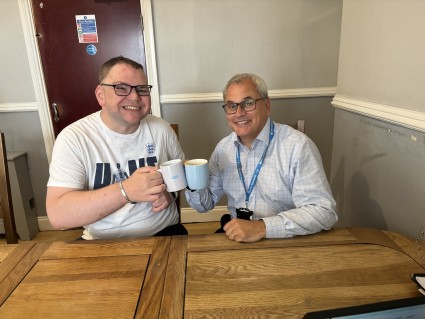 Two men sat at a dining table with cups of tea mugs and cheersing their mugs together